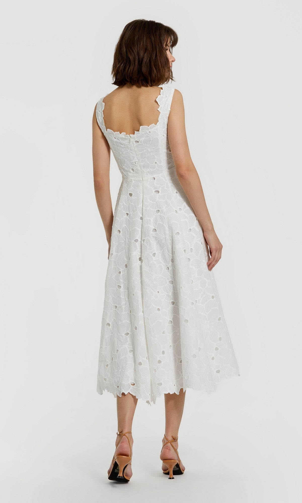 A woman stands facing away, displaying the elegant back of the Midi Party Dress: Mac Duggal 69096โa sleeveless white floral midi party dress adorned with delicate lace and eyelet-embroidered detailsโpaired with open-toe heels.